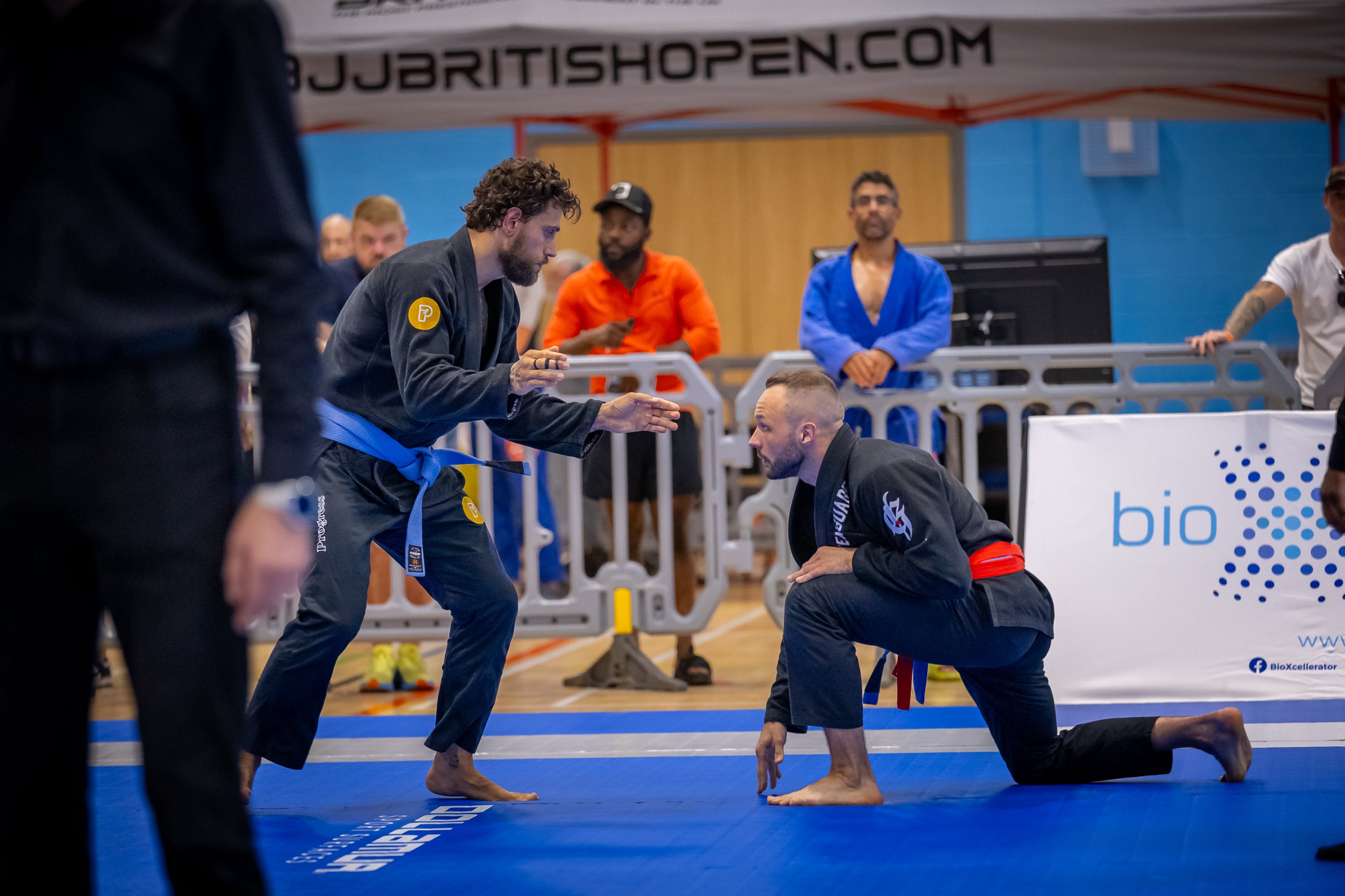 Two men in a Brazilian Jiu-Jitsu match at a competition with spectators and banners in the background.